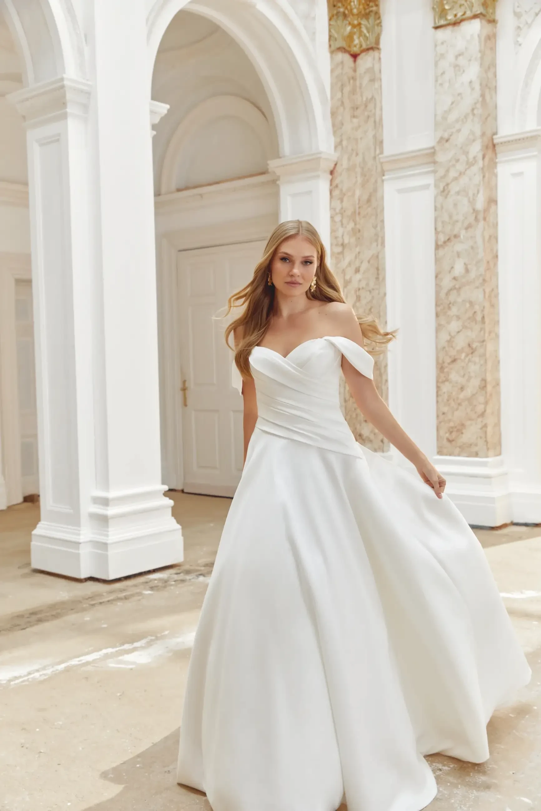 A woman wearing a white off-the-shoulder wedding dress is standing outdoors, with architectural columns and decorative walls in the background.