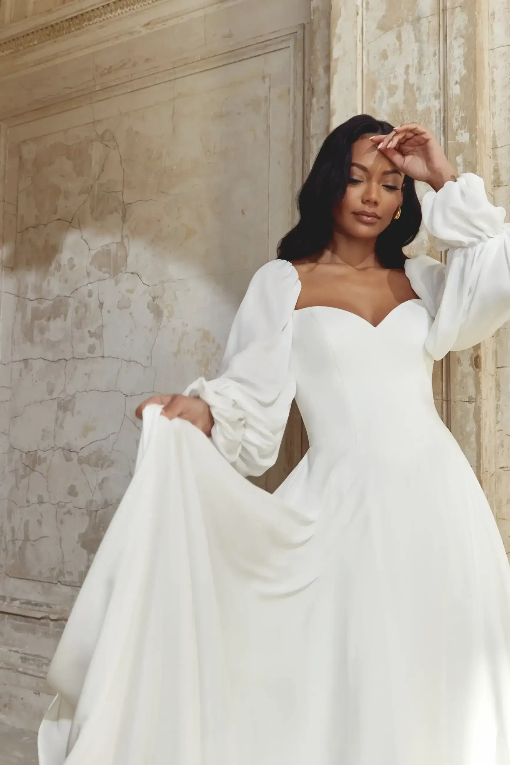 Woman in elegant white wedding dress with puffy sleeves poses gracefully against a textured, aged stone wall, conveying a serene and romantic ambiance.