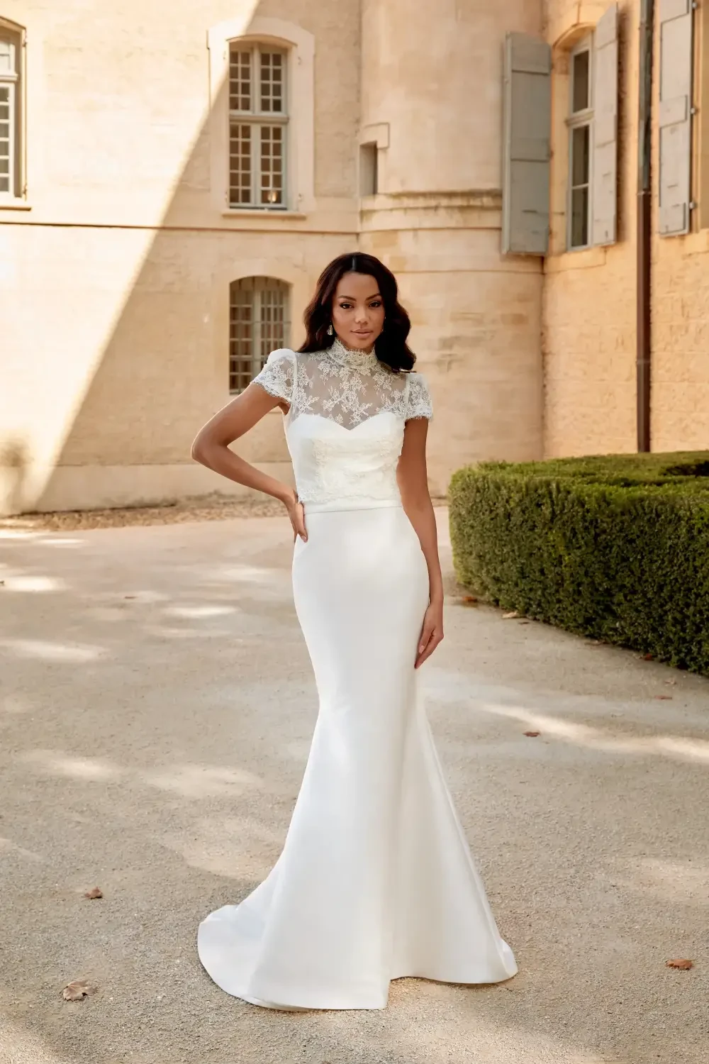 A woman wearing a fitted white wedding dress with lace details, standing outdoors in front of a building.