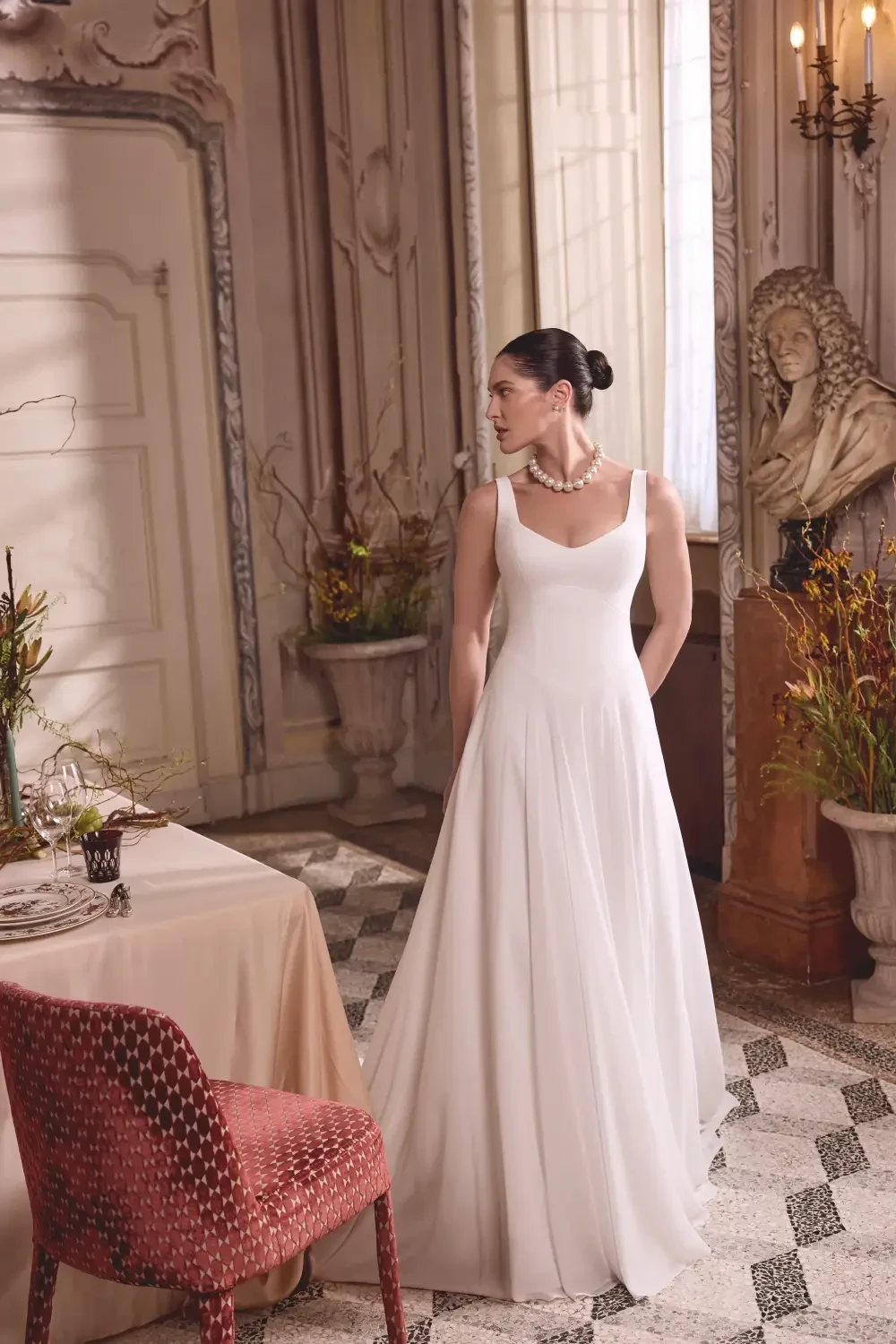 A woman in a white wedding dress stands elegantly in a tastefully decorated room, featuring classical decor and a statue in the background.
