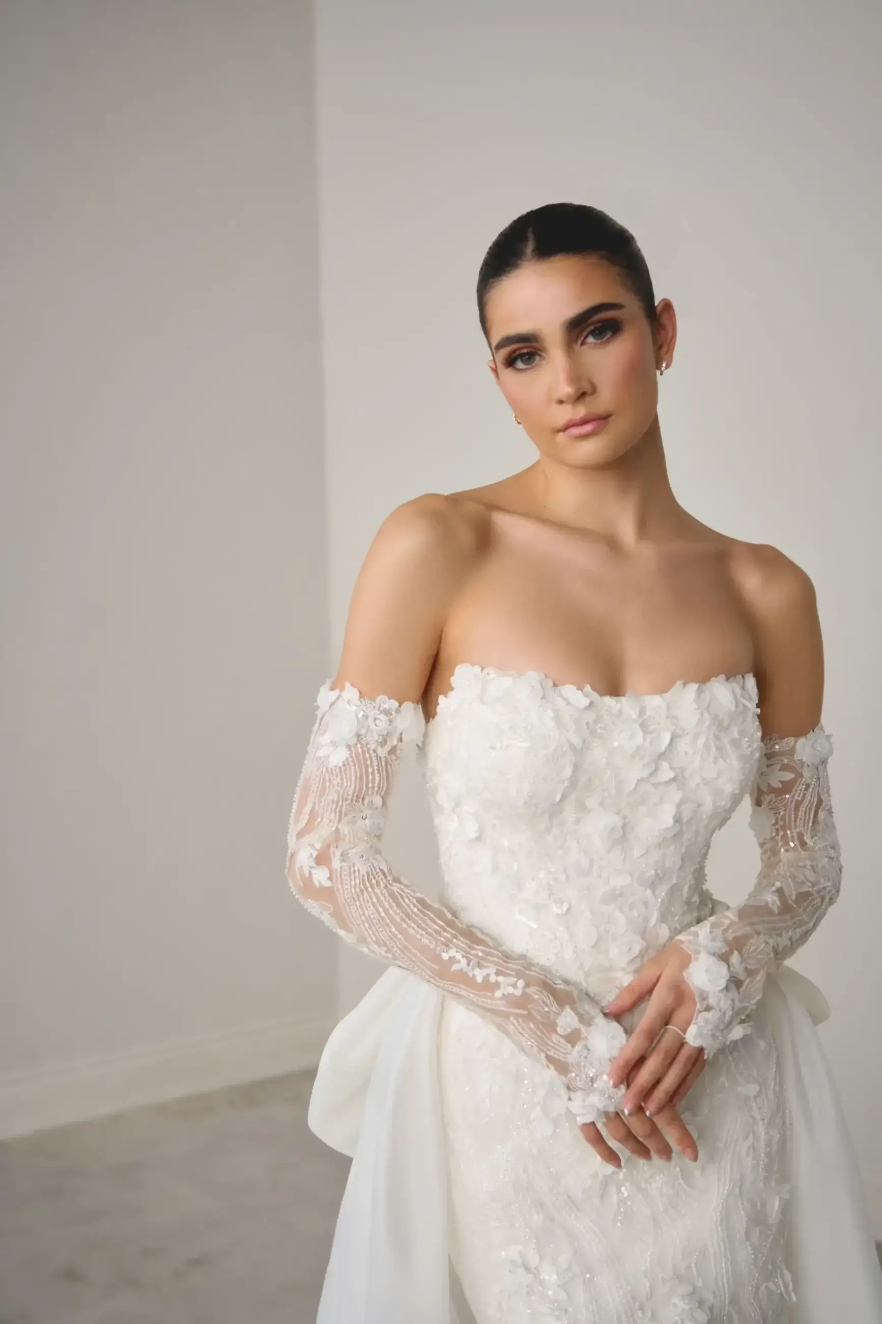 A woman in an elegant strapless white wedding dress adorned with floral lace stands against a plain backdrop. Her expression is serene and confident.