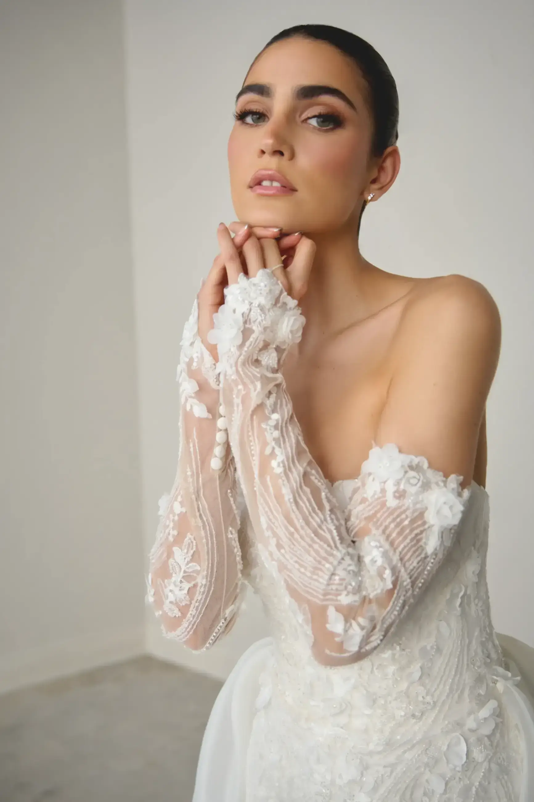 A woman in an elegant, off-shoulder white lace dress poses gracefully against a neutral background. Her expression is serene and confident.