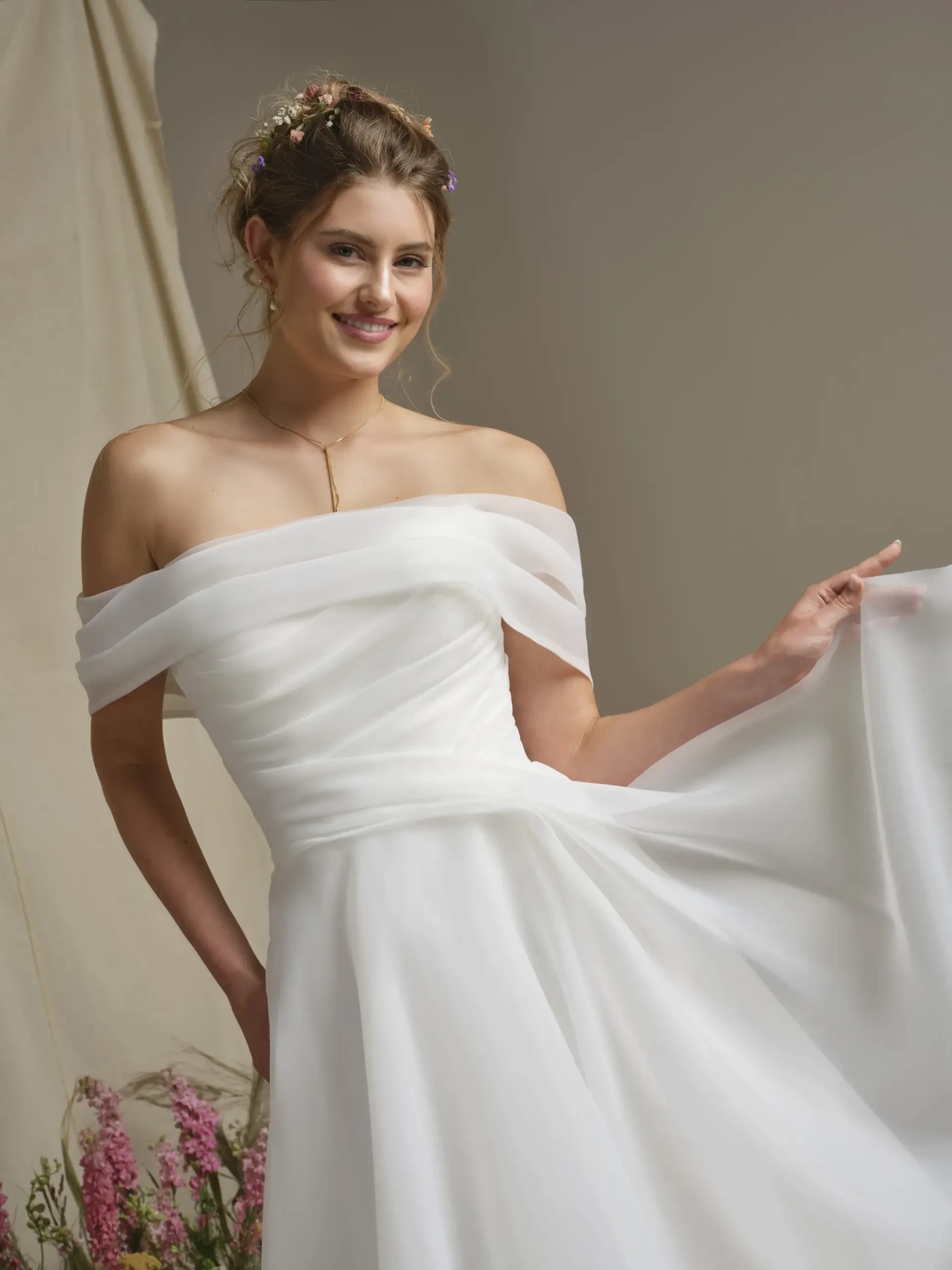 A young woman in an off-the-shoulder wedding dress smiles while holding the flowing fabric of her gown. She has a floral hair accessory and stands against a neutral background with soft lighting.
