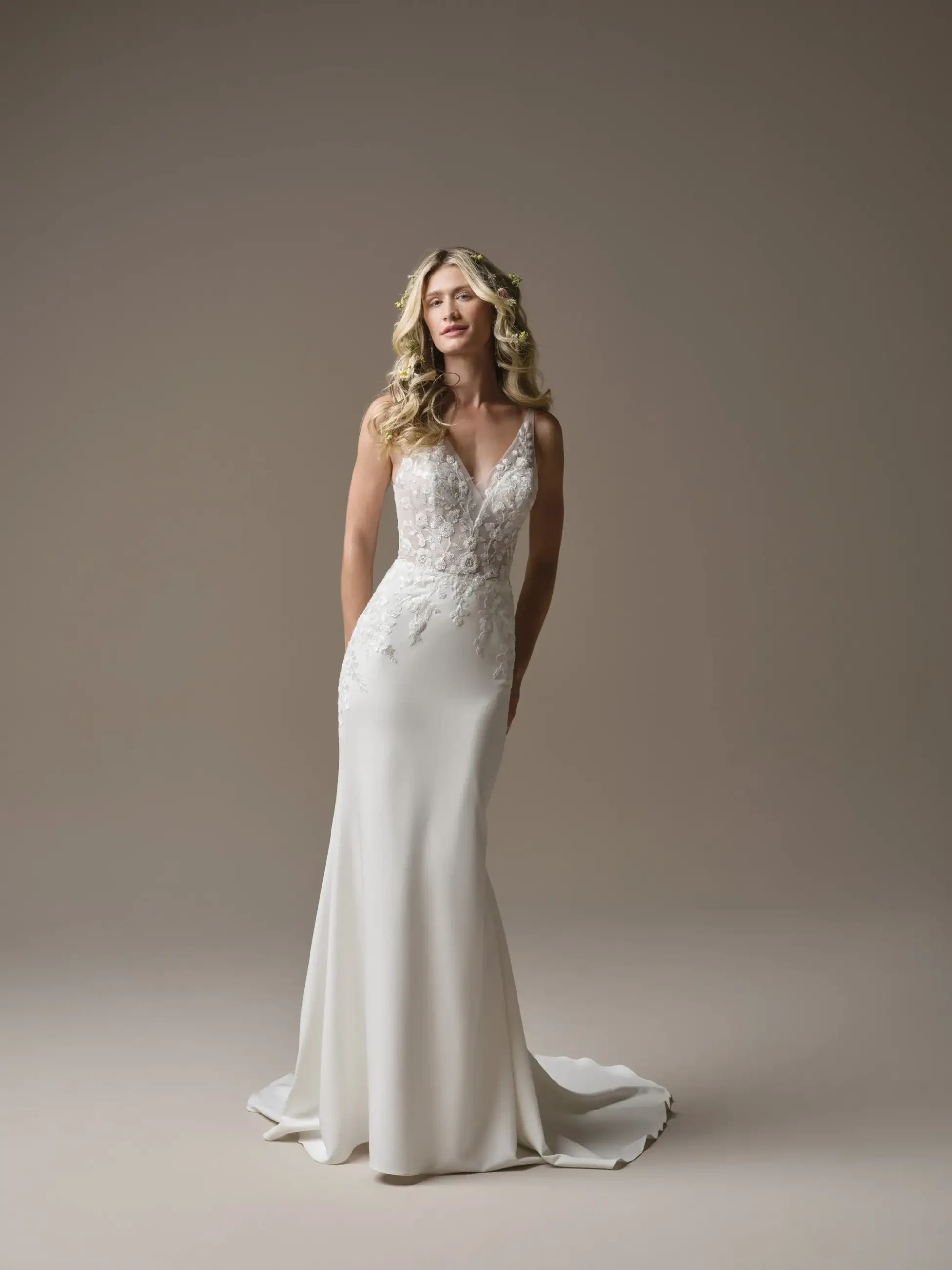 A woman in an elegant white wedding gown with lace details stands gracefully. Her long, wavy hair frames a serene expression. Neutral backdrop enhances the gown.
