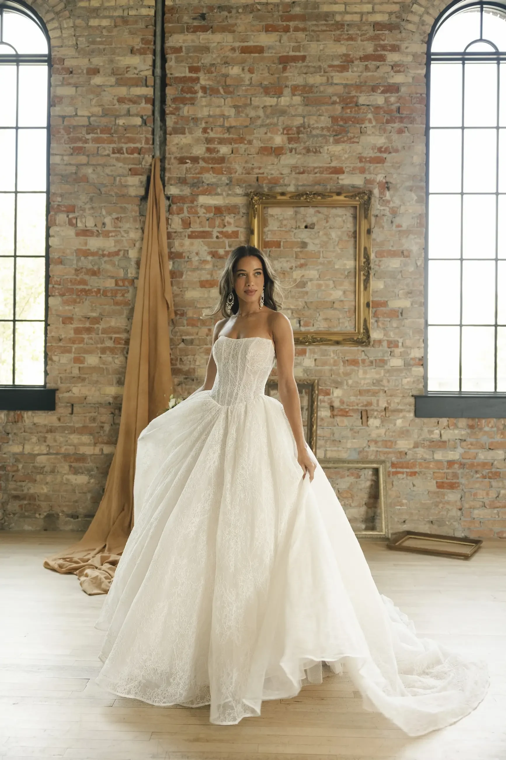 Bride in a flowing lace wedding gown stands in a bright, rustic room with exposed brick and tall windows, evoking elegance and romance.