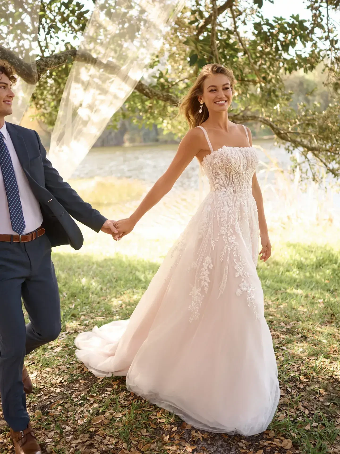 A bride in a beautiful wedding dress holding hands with a man, smiling while walking outdoors near a lake, surrounded by trees.