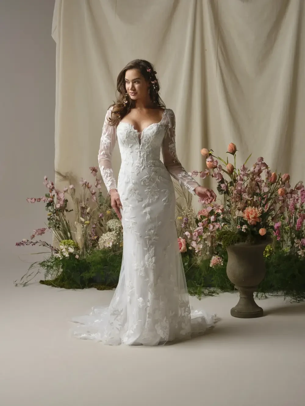 A woman wearing a long-sleeved wedding dress stands in front of a floral arrangement, smiling gracefully.