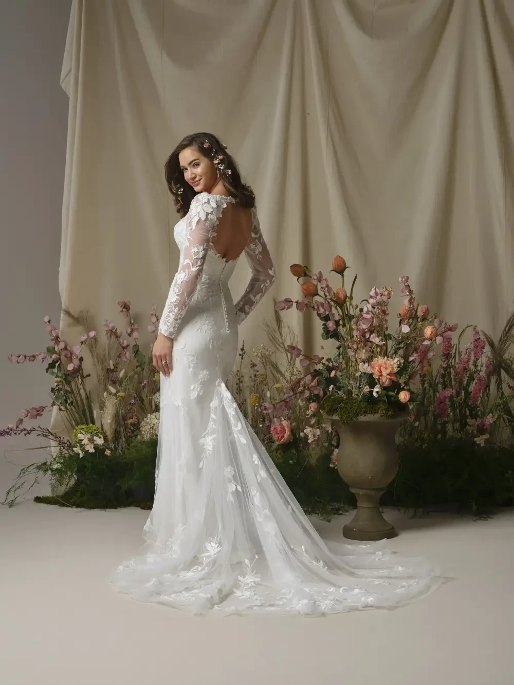 A bride in a white wedding dress with long sleeves and floral lace detailing, standing with her back to the camera, surrounded by a floral arrangement and a neutral backdrop.