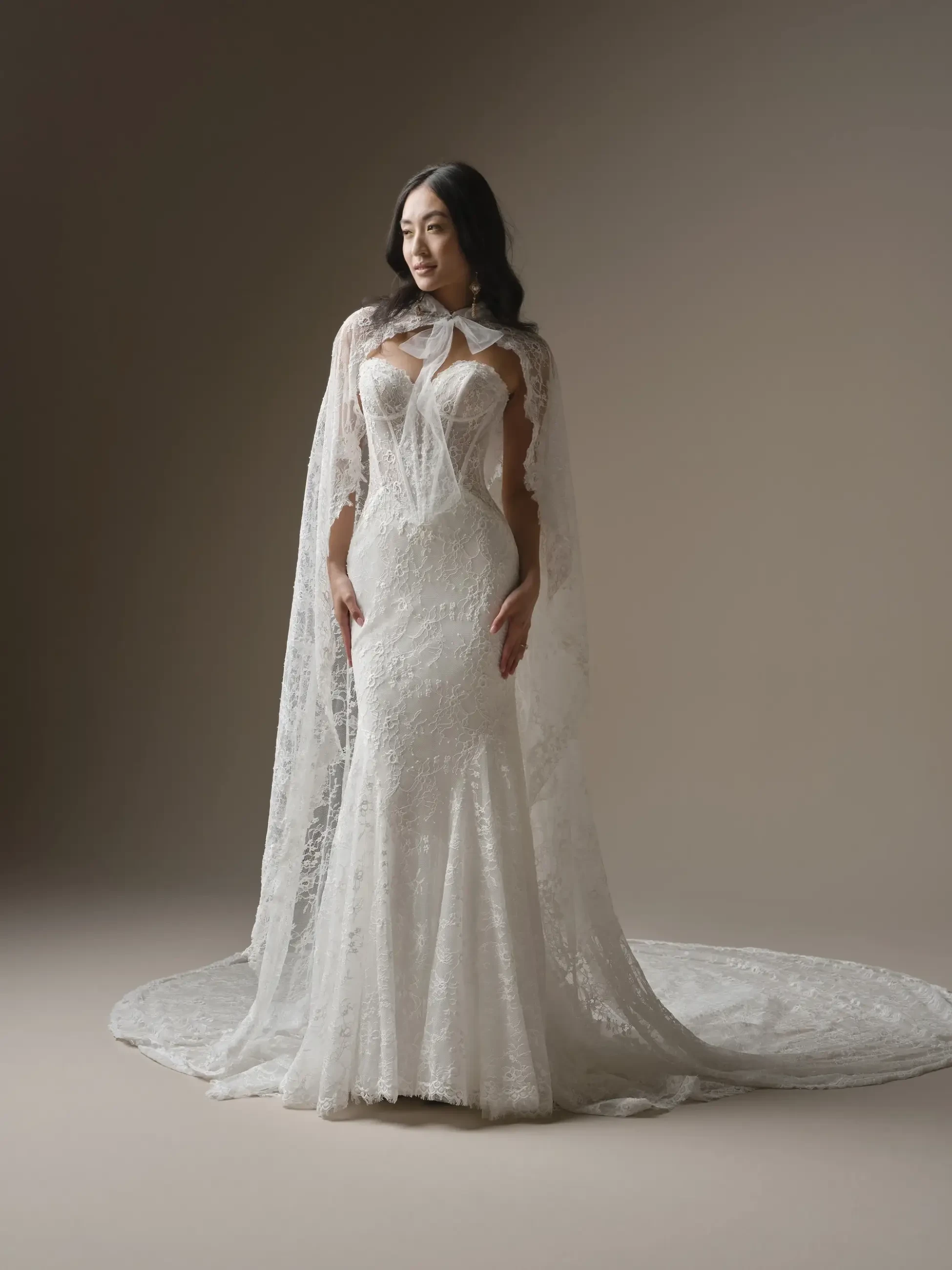 A woman in an elegant, lace wedding gown with a long, flowing cape stands against a neutral background, exuding a serene and confident presence.
