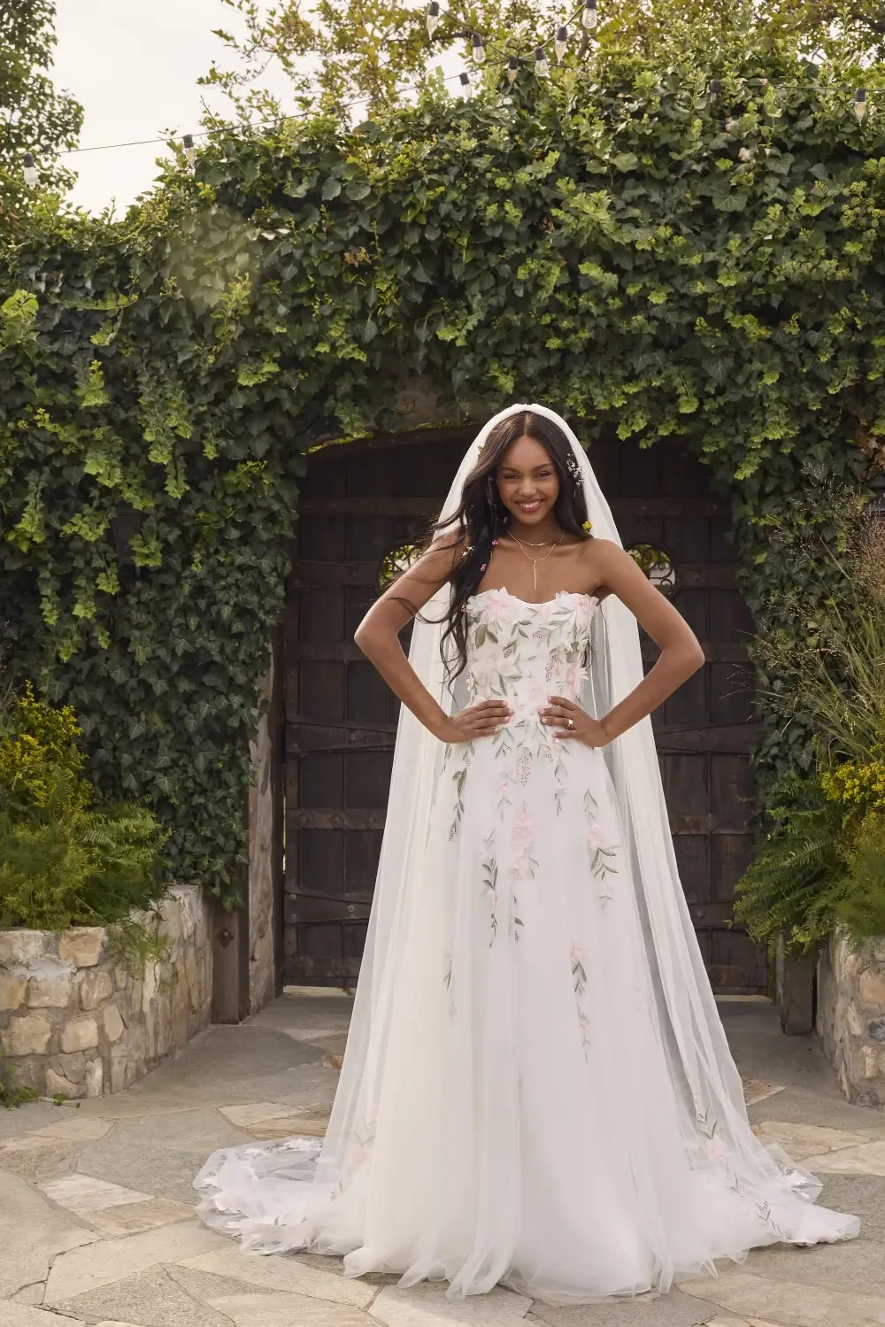 A bride in a strapless wedding gown with floral embroidery, standing in front of a rustic wooden door surrounded by greenery. She has long hair and is wearing a veil.