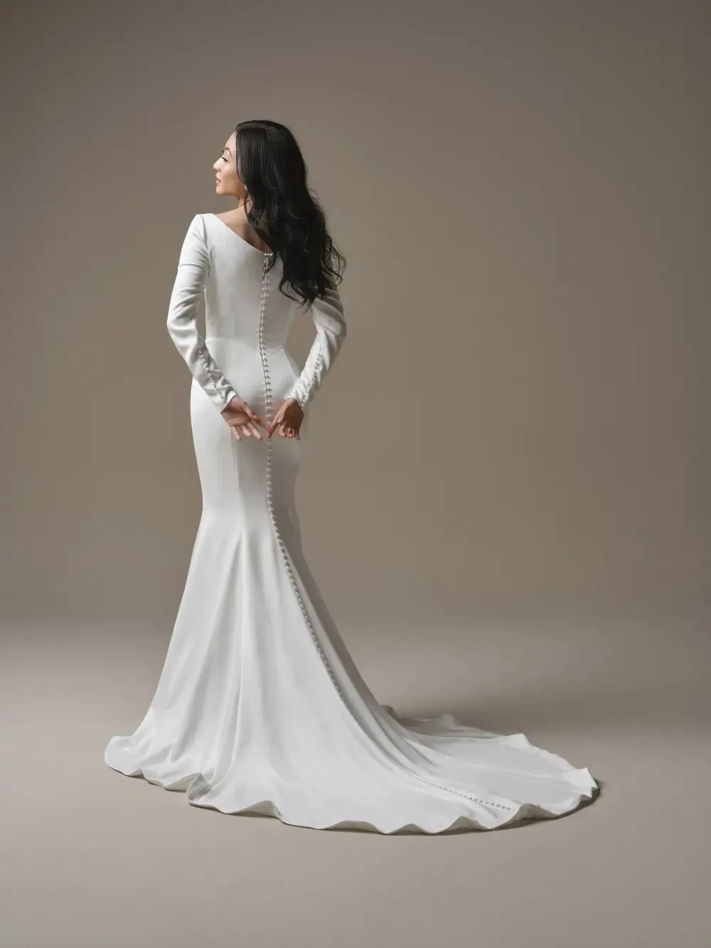 A woman in a long, form-fitting white dress with a train, standing with her back to the camera, her hair cascading down her shoulders. The dress features button detailing along the spine and long sleeves.